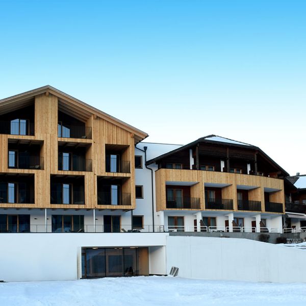 Modern Alpine hotel exterior in winter with contemporary wooden facade, snow-covered grounds, and mountain backdrop under blue sky