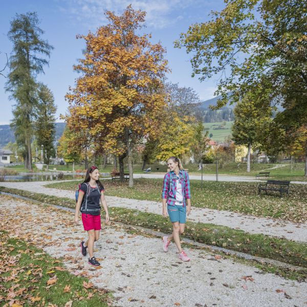 Two women hiking on autumn path through park with golden trees and mountains in background