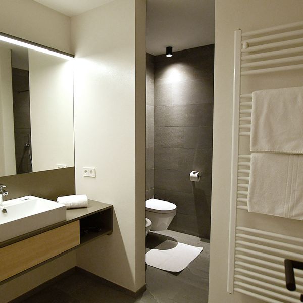 Modern bathroom with floating wooden vanity, large backlit mirror, LED strip lighting, and separate toilet area with dark tile accent wall