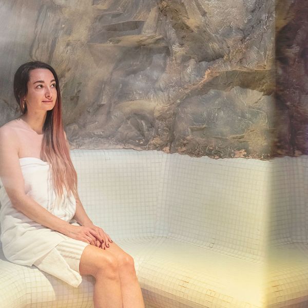 Woman in white towel relaxing in spa steam room with textured stone wall features and soft ambient lighting
