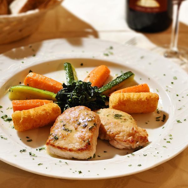 Elegant plated fish dish with glazed vegetables, golden potato croquettes, and herb sauce on white plate with wine