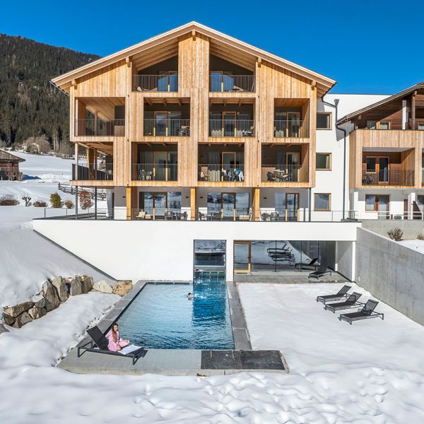 Modern Alpine hotel exterior in winter with heated outdoor pool, wooden balconies, and snow-covered mountain backdrop under blue sky