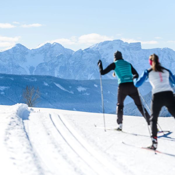 Two cross-country skiers on groomed snow trail with dramatic panoramic view of Dolomite mountain peaks