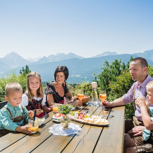 Family in traditional dirndls and lederhosen enjoying outdoor dining on mountain terrace with dramatic Dolomite peaks backdrop