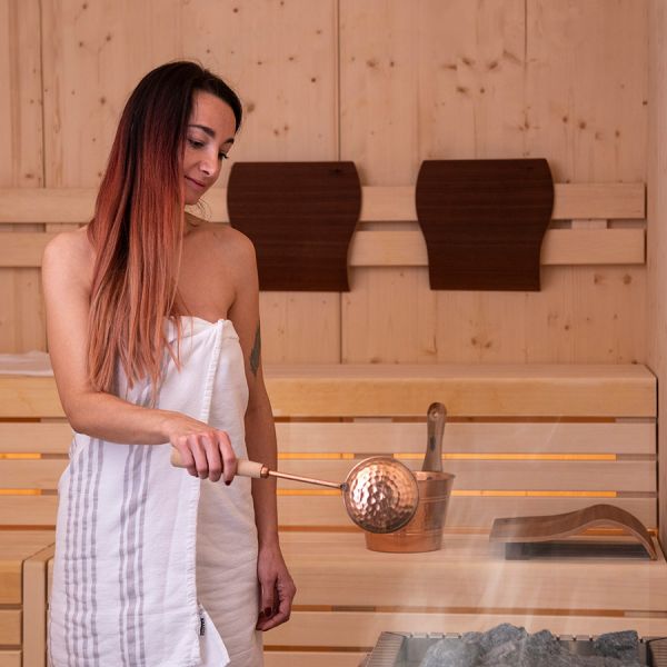 Woman pouring water on sauna stones with copper ladle in modern wooden sauna with LED-lit benches