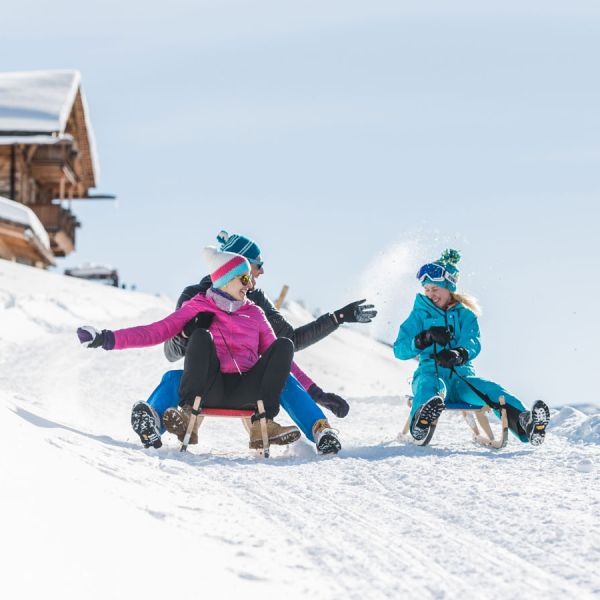 Three people on wooden sleds racing down snowy slope creating snow spray with traditional Alpine chalet in background