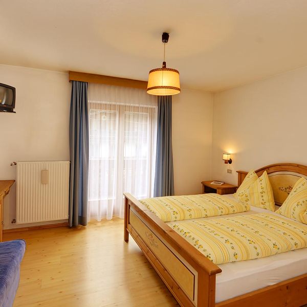 Traditional hotel bedroom with pine wood furniture, yellow patterned bedding, wall-mounted TV, and natural lighting