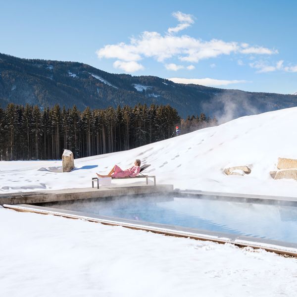 Woman relaxing on lounge chair beside steaming heated outdoor pool surrounded by pristine snow with forested mountain view