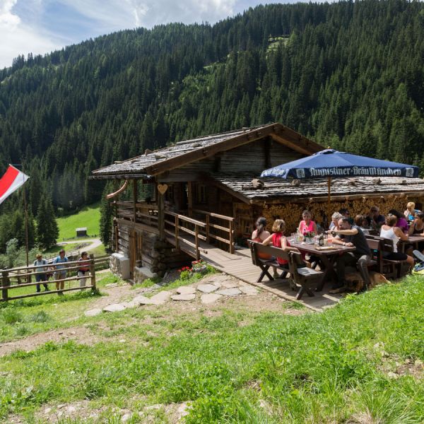 Traditional Alpine mountain hut restaurant with outdoor terrace dining, Austrian flag, and dense forest-covered slopes backdrop