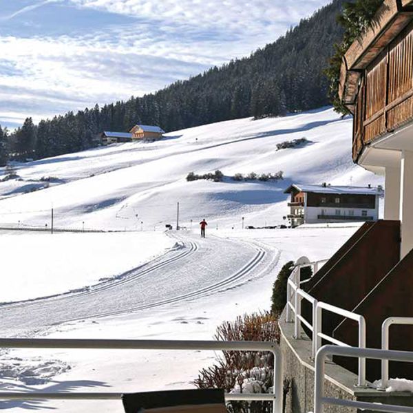 Winter view from hotel terrace of groomed cross-country ski trail through snowy valley with traditional Alpine chalets and mountains