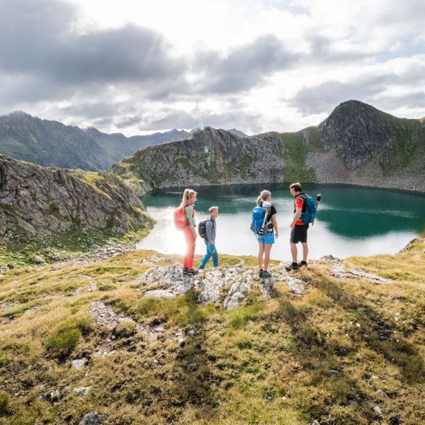 Four hikers with backpacks standing on rocky mountain ridge overlooking turquoise glacial lake surrounded by dramatic peaks
