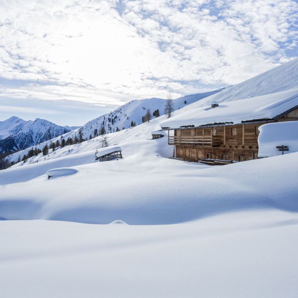 Traditional rustic Alpine wooden cabin heavily buried in deep pristine snow on mountain slope with ski area backdrop