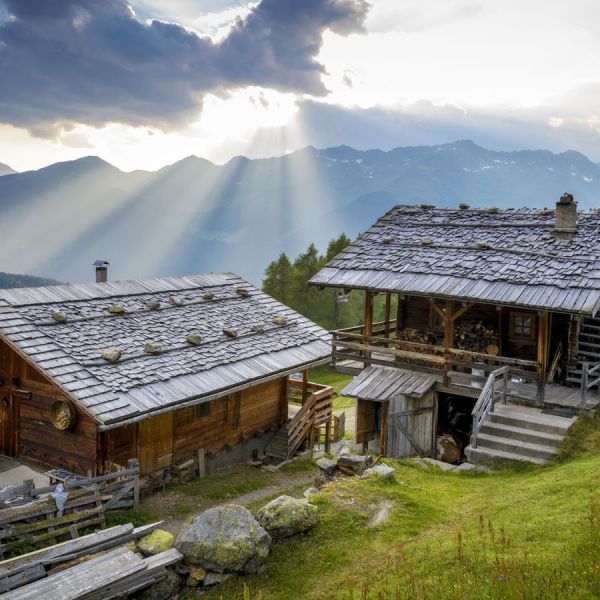 Two traditional rustic wooden Alpine mountain chalets on green hillside with dramatic sunset light rays breaking through storm clouds