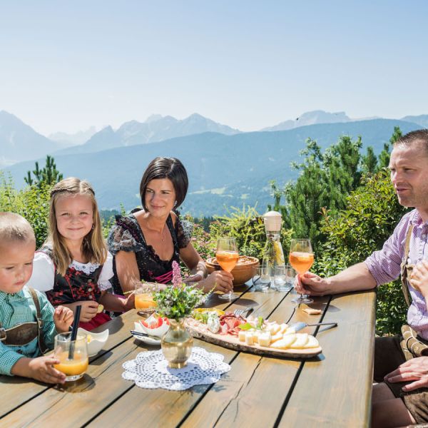 Family of five in traditional dirndls and lederhosen enjoying drinks and cheese platter on mountain terrace with Dolomite peaks panorama
