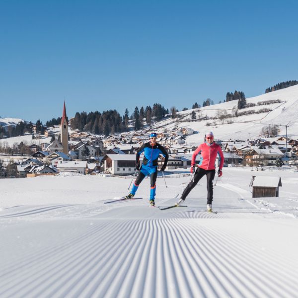 Two cross-country skiers in bright sportswear on perfectly groomed snow trail with Alpine village and blue sky backdrop
