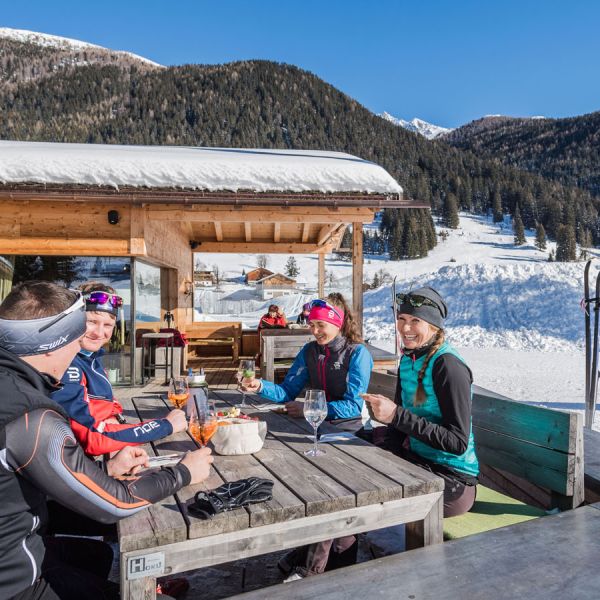 Group of cross-country skiers in colorful gear relaxing with drinks at sunny mountain hut terrace with snowy peaks view
