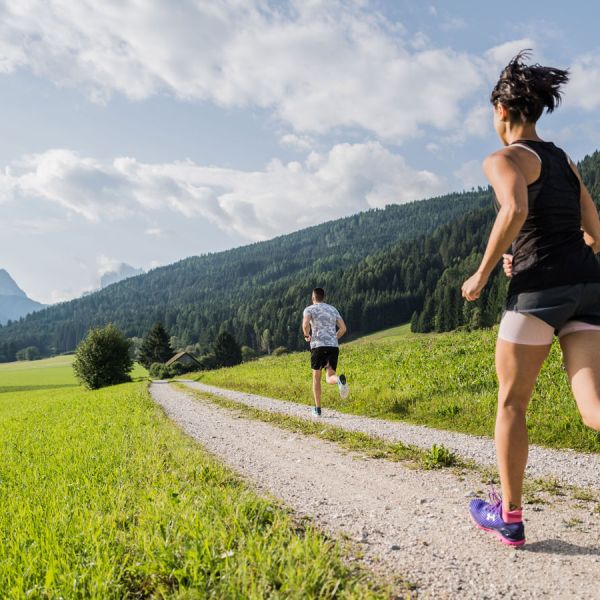 Two runners jogging on rural gravel road through vibrant green Alpine meadows with forested mountain slopes and clouds
