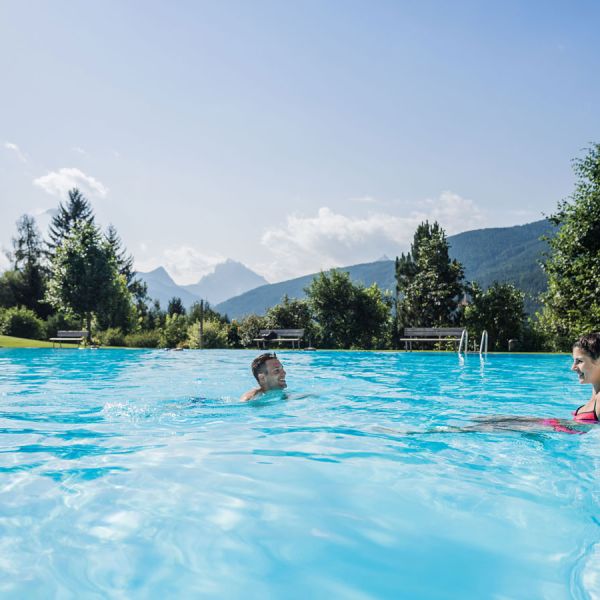 Couple swimming and relaxing at edge of large outdoor pool surrounded by mountain forest landscape under blue sky